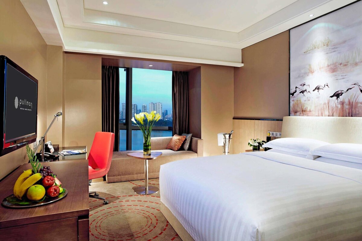 A hotel room with a bed, a desk with a TV, a chair, a small table with a vase, and a painting on the wall.