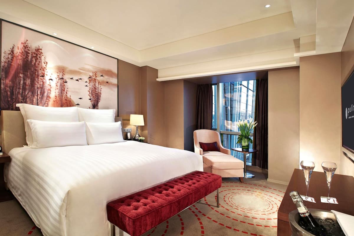 Habitación de hotel con una cama grande, un banco rojo, una mesita con vasos y vistas a la ciudad a través de amplias ventanas.