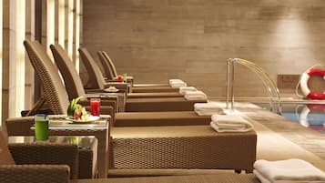 A row of wicker lounge chairs with towels and a glass of green drink, a pool area with a lifebuoy, and a tiled wall in the background.