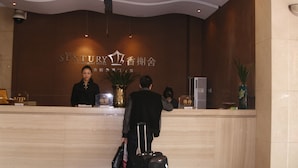Un mostrador de recepción de hotel con un letrero que dice "Sentury Apartment Hotel". Hay dos recepcionistas detrás del mostrador, y un hombre con una maleta está de pie frente a él.