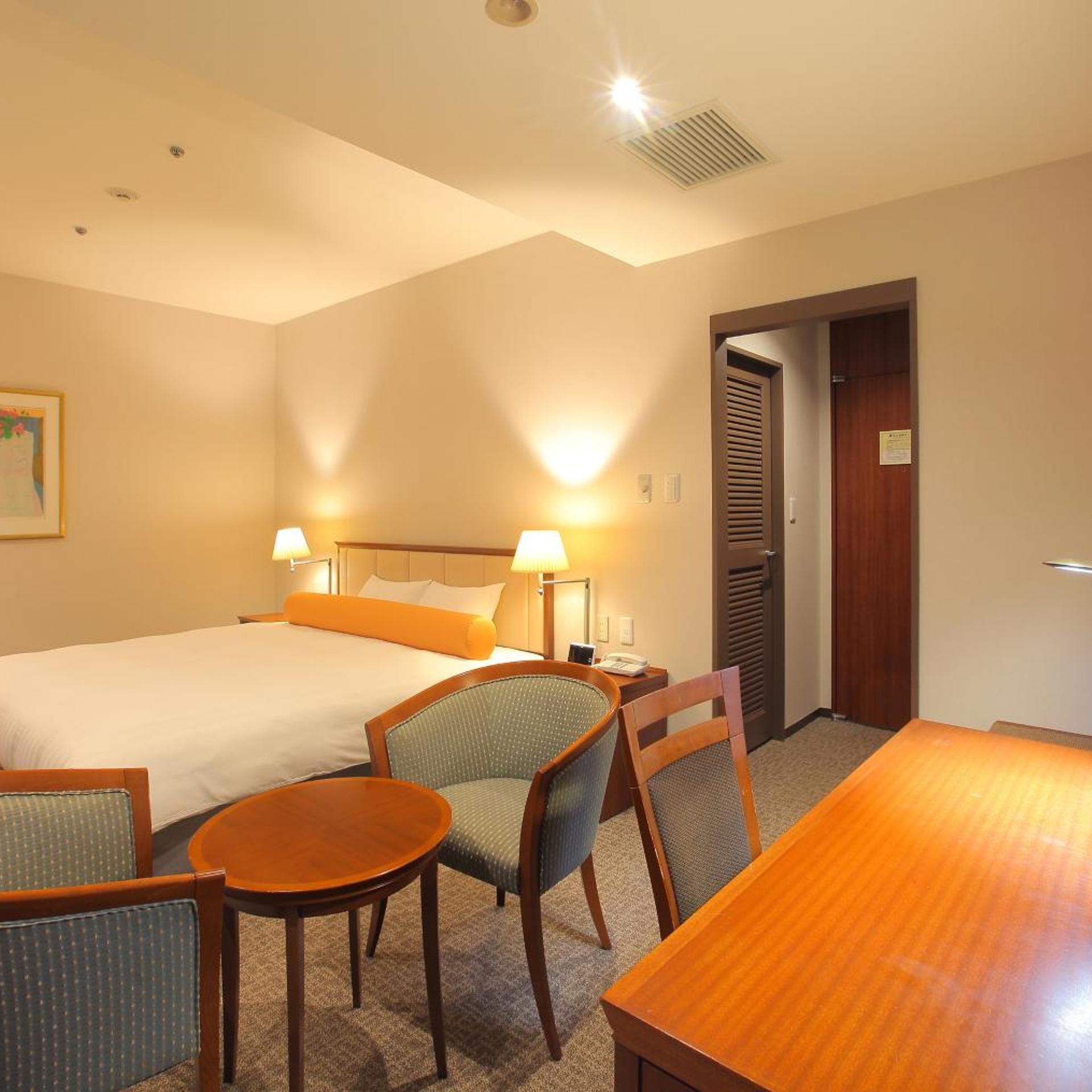 A hotel room with a bed, two bedside lamps, a chair, a small table, and a door.
