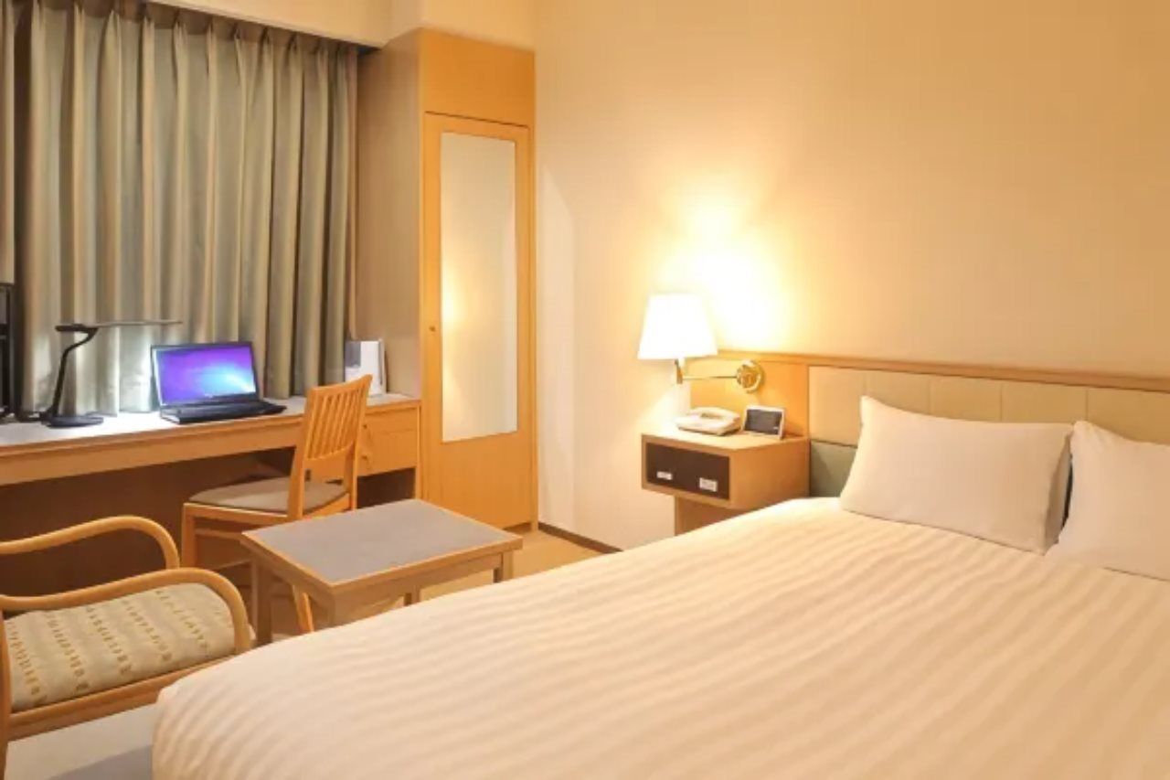 A hotel room with a bed, a desk with a laptop, a chair, a nightstand with a lamp, and a window with curtains.