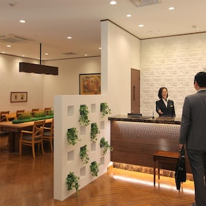 Zona de recepción con un mostrador de madera, un área de espera con una mesa alargada y sillas, y una pared con un diseño geométrico.