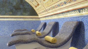A room with mosaic-tiled walls and ceiling, featuring a large arched window and a seating area with towels.