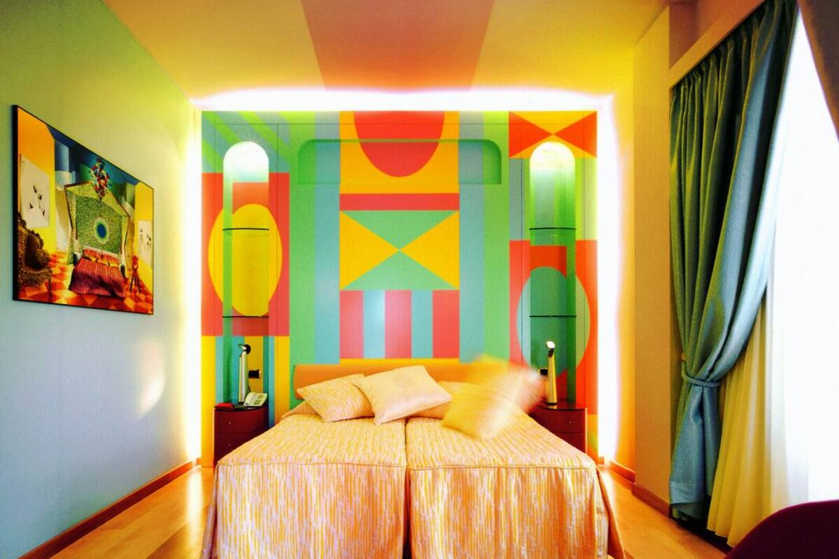 A brightly colored room with a large bed, a painting on the wall, and a window with curtains.