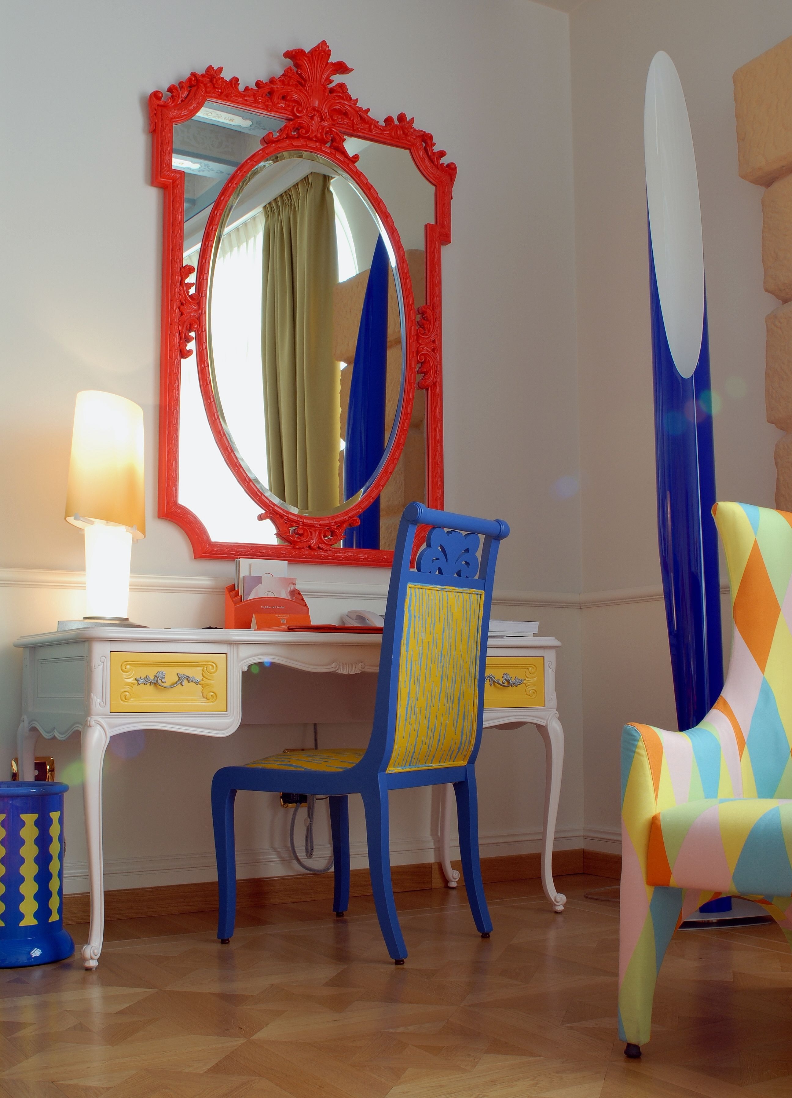 Una stanza con una scrivania bianca, una sedia blu, una poltrona colorata e uno specchio con cornice rossa.