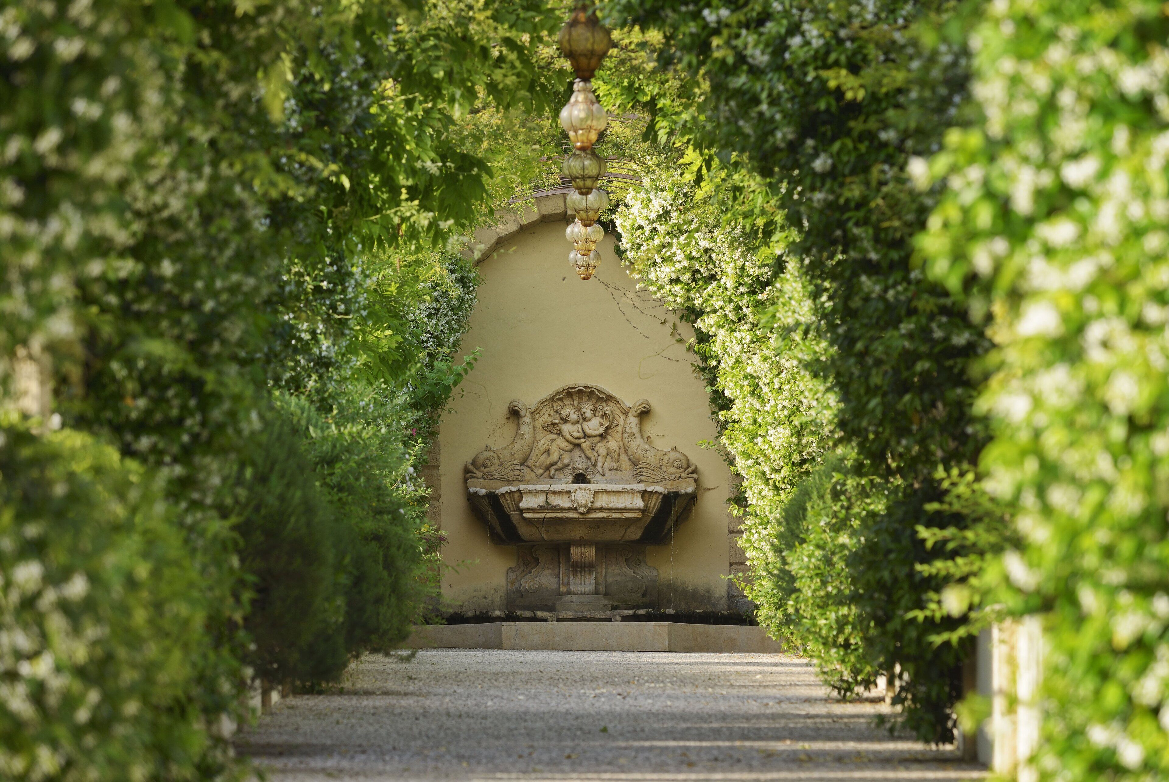 A stone pathway leading to a fountain with a decorative wall and greenery on either side.