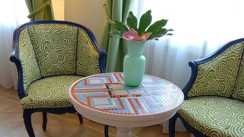 Una habitación con dos sillas estampadas, una mesa redonda con cubierta de mosaico y un jarrón verde con una flor rosa.