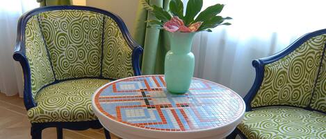 Una habitación con dos sillas estampadas, una mesa redonda con cubierta de mosaico y un jarrón verde con una flor rosa.