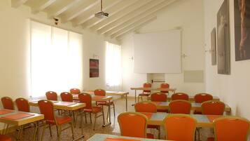 A classroom with rows of orange chairs and tables, white walls, and a ceiling with exposed beams.