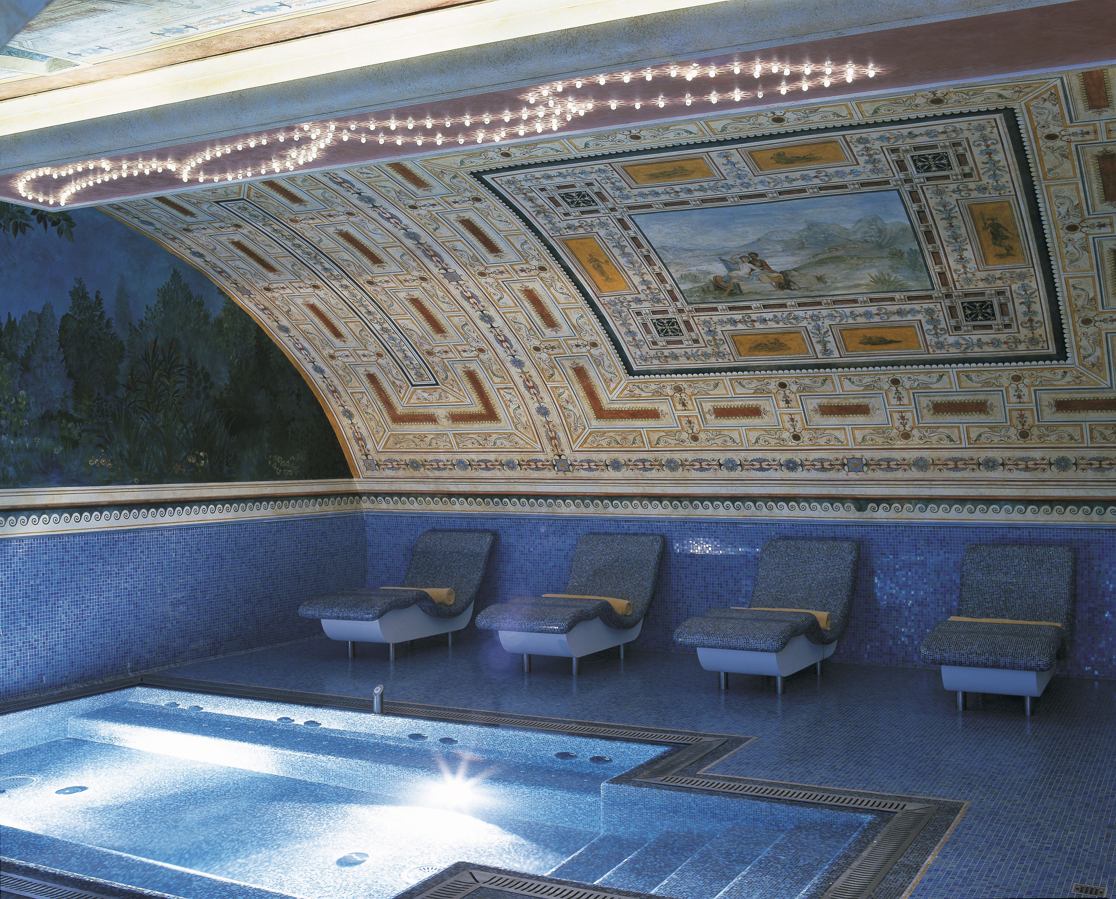 A luxurious indoor pool area with a tiled floor, a mural on the ceiling, and several seating areas.