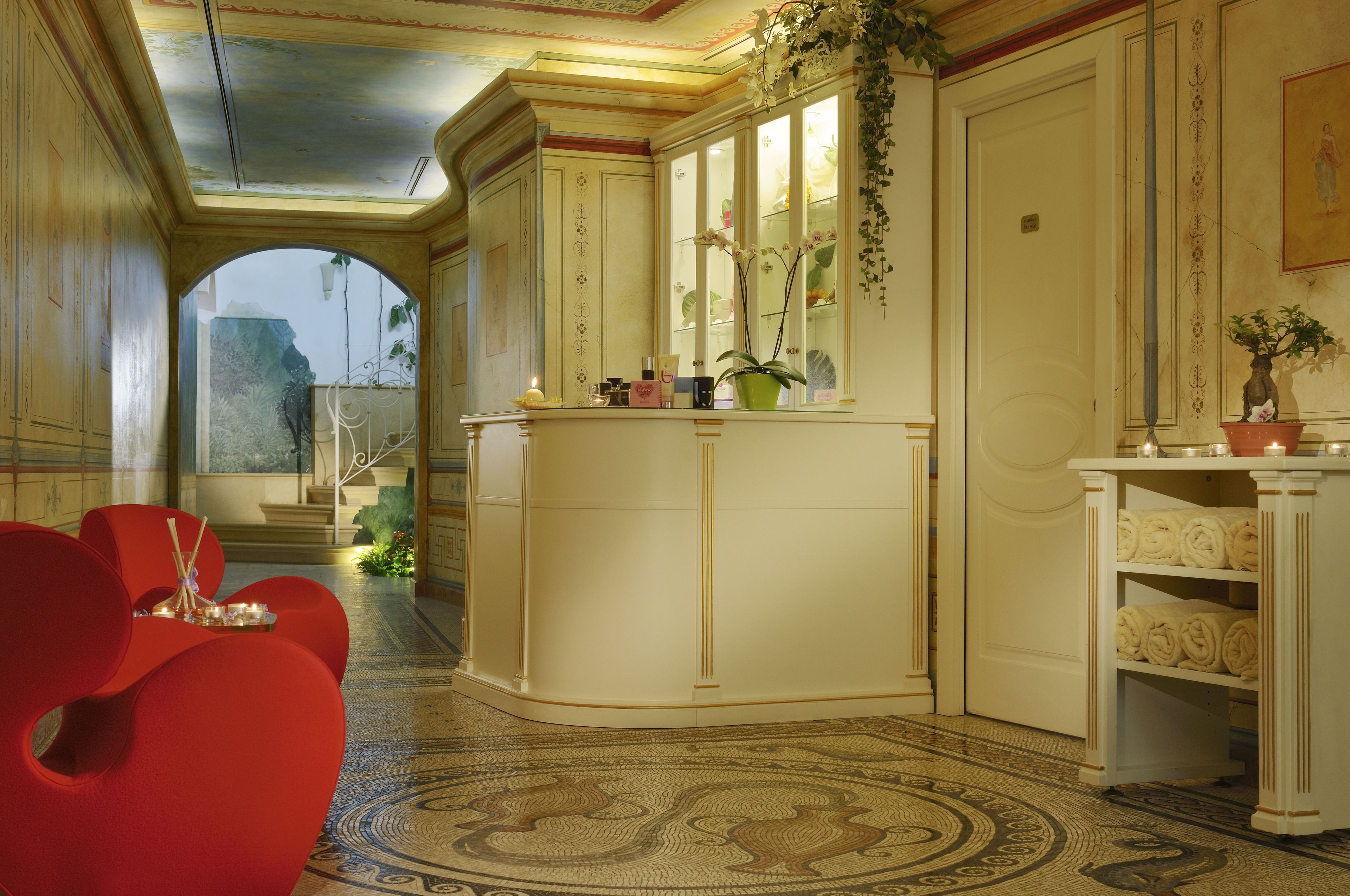 Um hall de hotel luxuoso, com um balcão de recepção curvo, área de estar em vermelho e piso estampado.