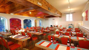 A dining area with multiple tables and chairs, wooden ceiling beams, and large windows.