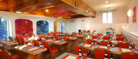A dining area with multiple tables and chairs, wooden ceiling beams, and large windows.