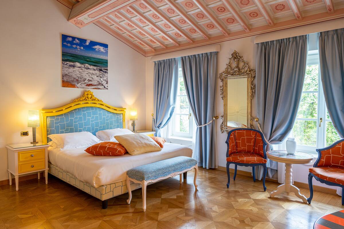A bedroom with a bed, bedside table, bench, two chairs, a round table, a mirror, and a painting of a beach scene.