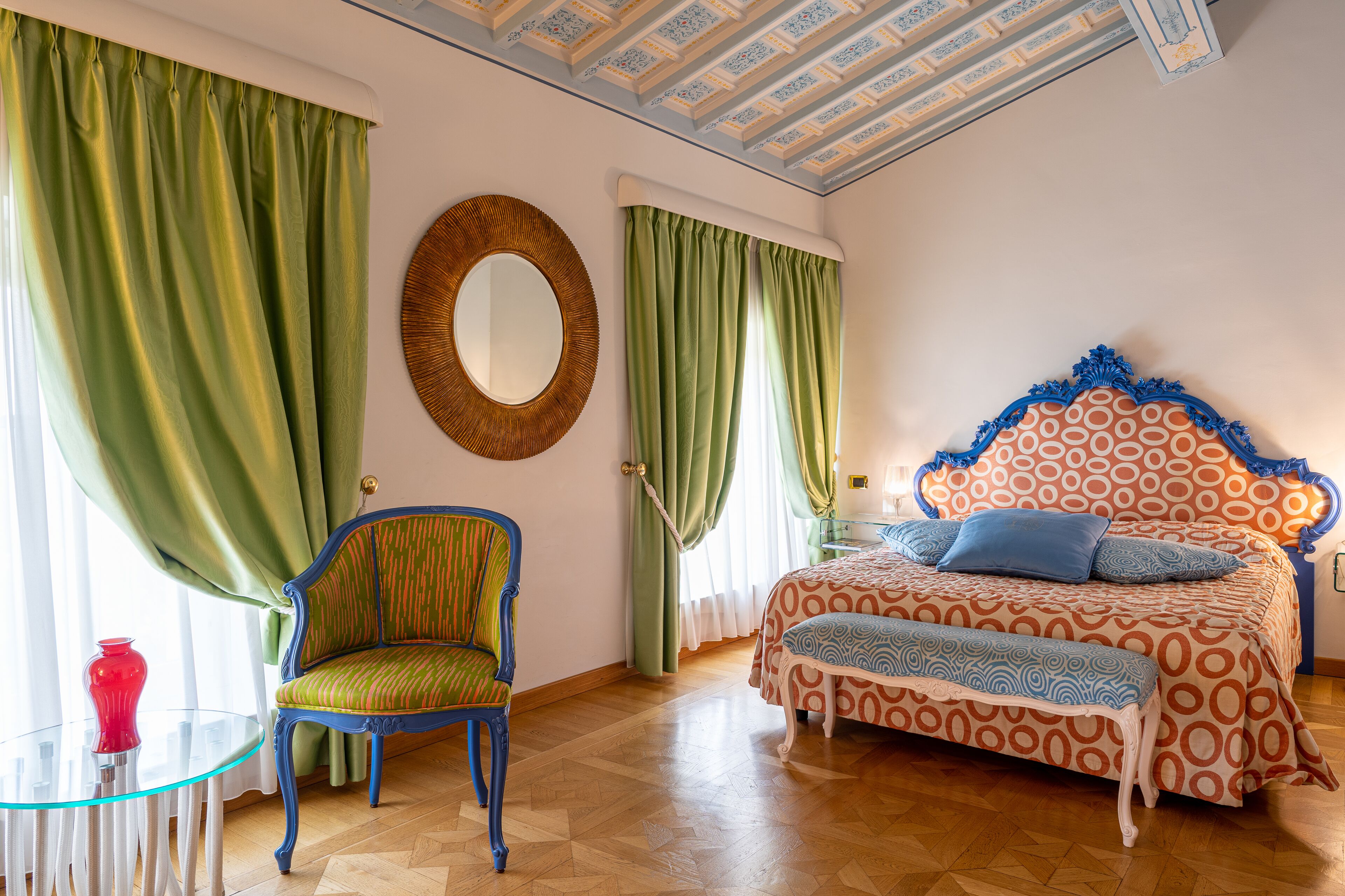 A bedroom with a bed, a chair, a round mirror, and a glass table.