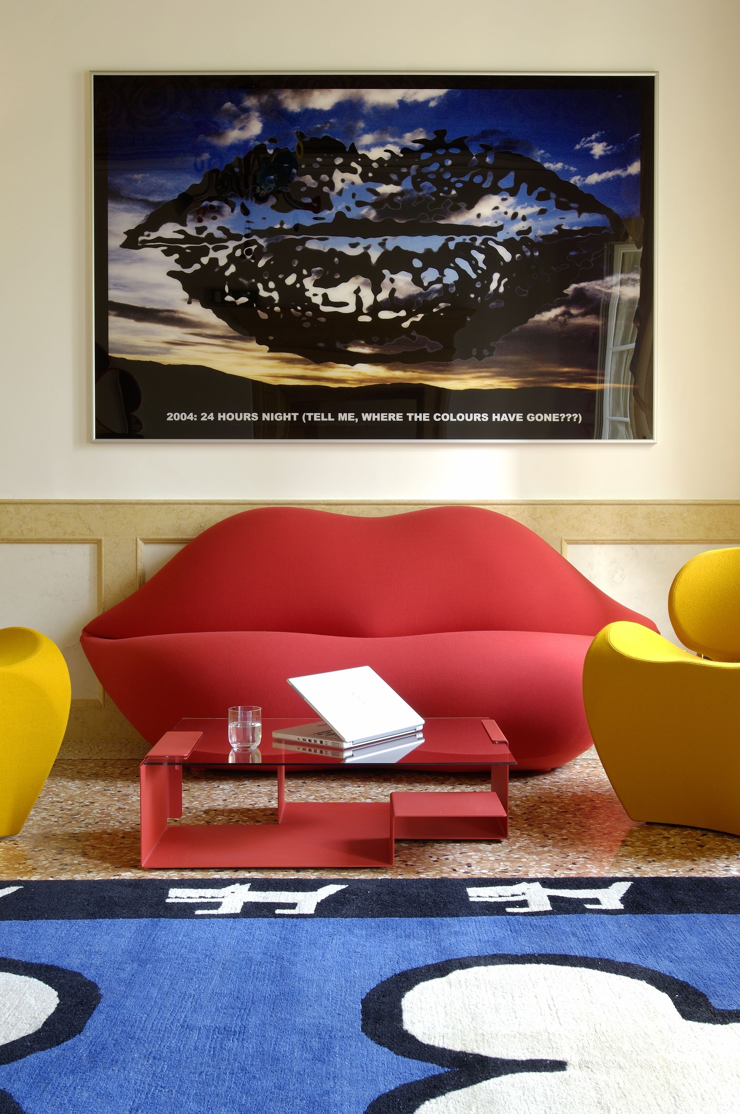 Een moderne woonkamer met een rode bank, een gele fauteuil en een klein rode salontafel met een laptop en glas.