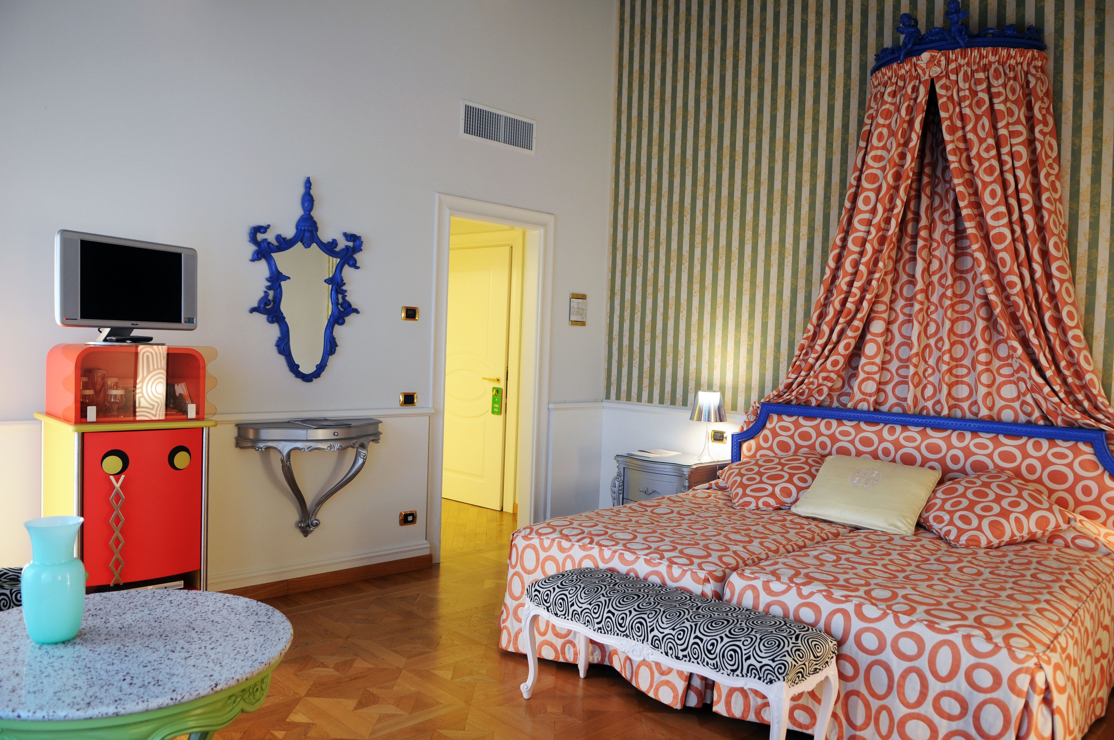 A bedroom with a four-poster bed, a television on a cabinet, a small table with a vase, and a striped curtain.