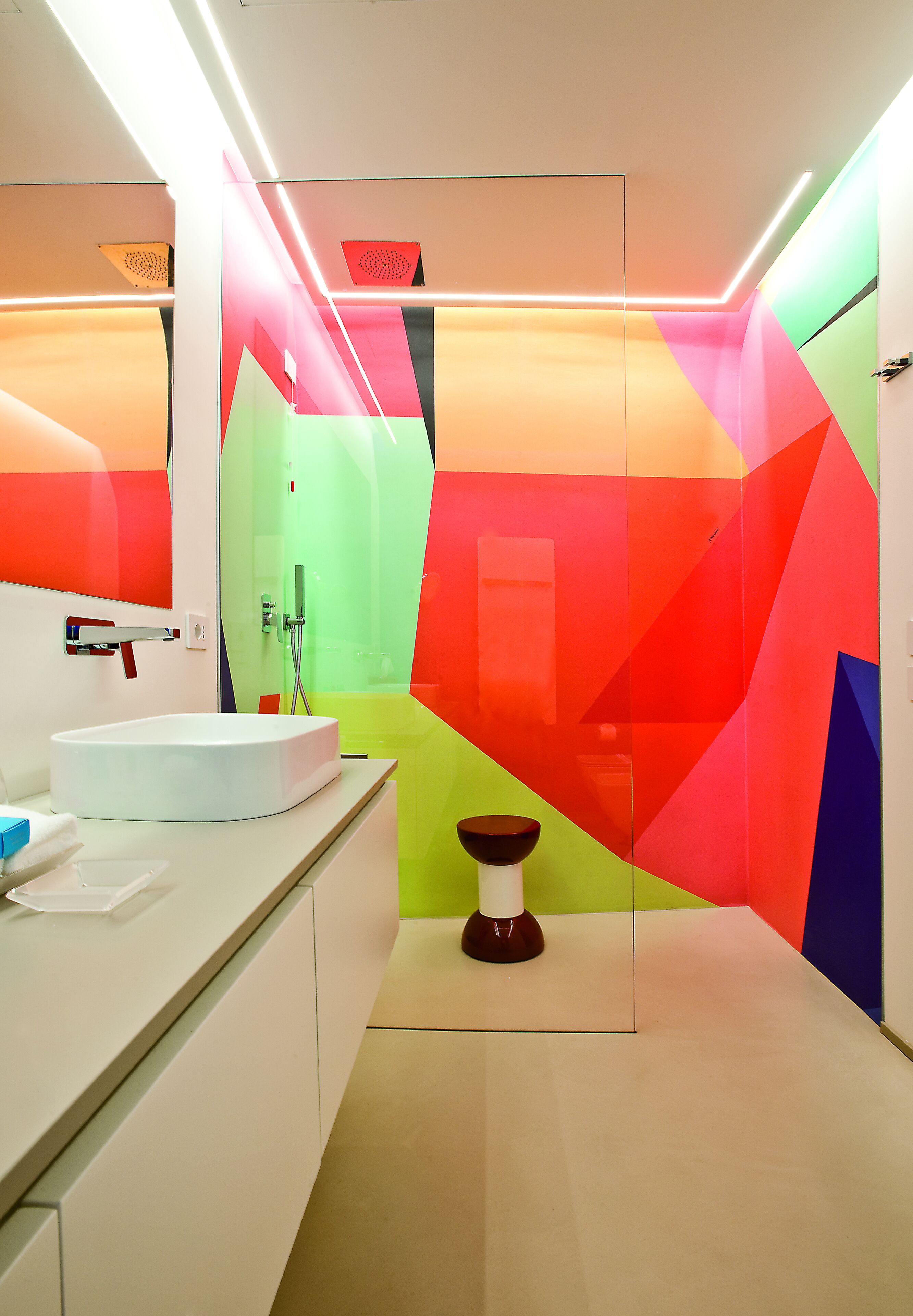 Ein modernes Badezimmer mit einem weißen Waschbecken, einer Duschkabine aus Glas und einem farbenfrohen, geometrischen Wanddesign.
