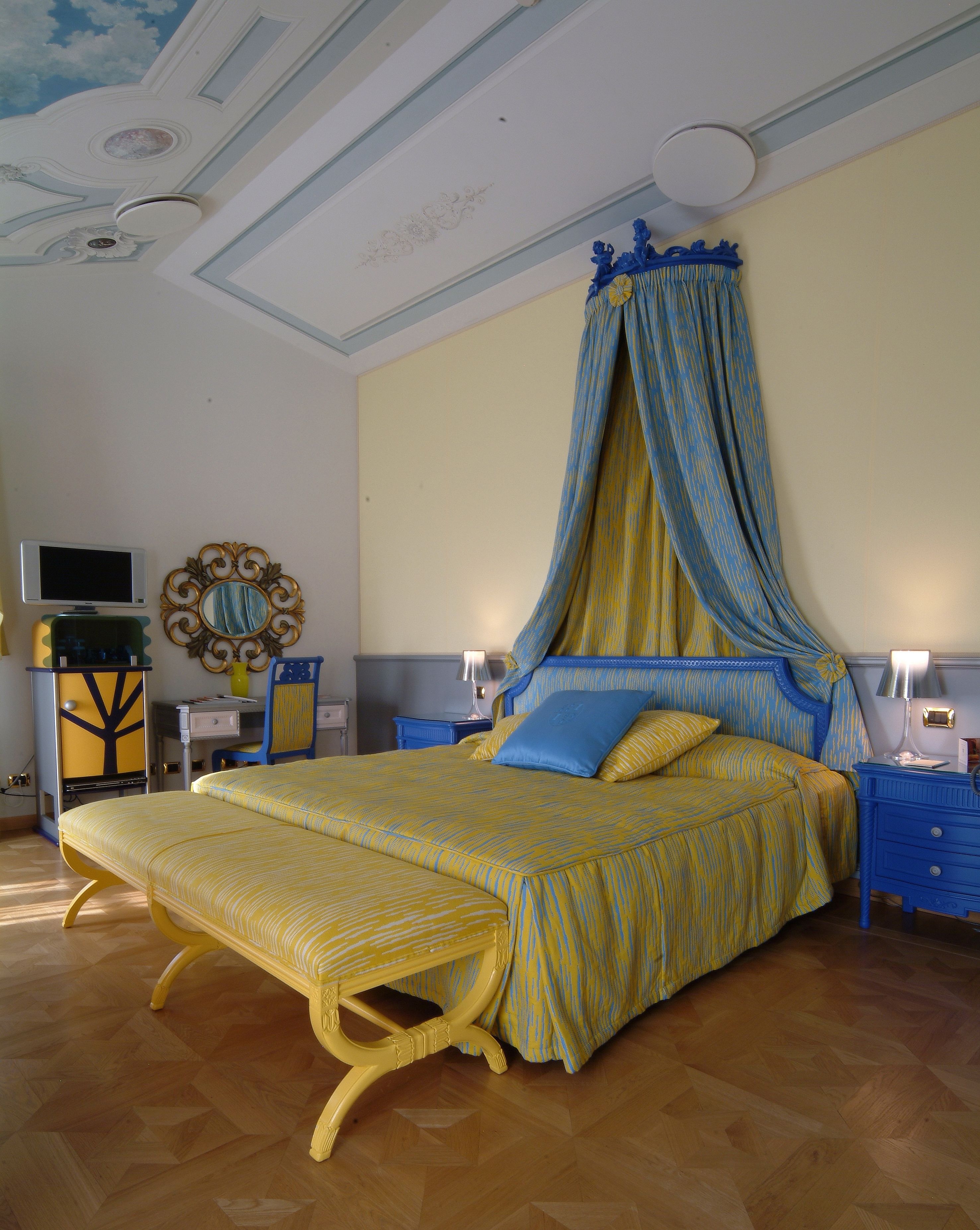 A bedroom with a four-poster bed, a television, a mirror, and a desk with a lamp.