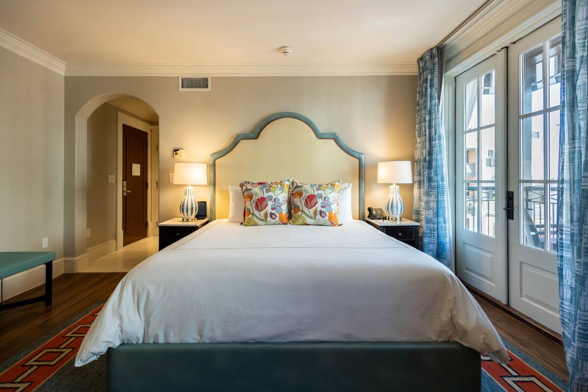 A hotel room with a large bed, two bedside tables with lamps, a bench, a door, and a window with curtains.
