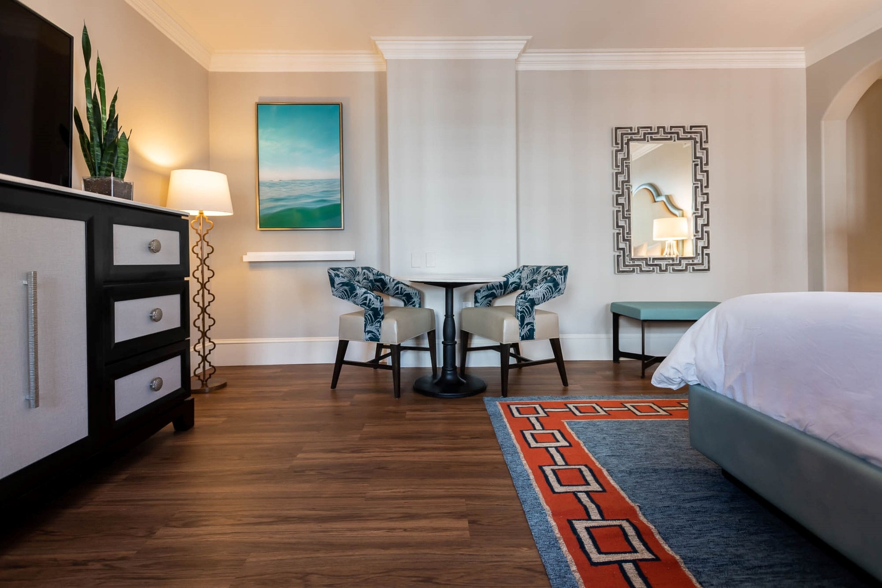Una habitación de hotel con una cama, un escritorio con dos sillas, una lámpara, una planta, un cuadro y un espejo.