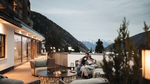 A cozy outdoor seating area with a fire feature, a glass balustrade, and a view of mountains. - Falkensteiner Hotel Falkensteinerhof  (Rio di Pusteria)