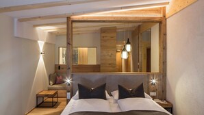 A bedroom with a large bed, a wooden headboard, a nightstand, a bench, and a mirror. - Falkensteiner Hotel Falkensteinerhof  (Rio di Pusteria)
