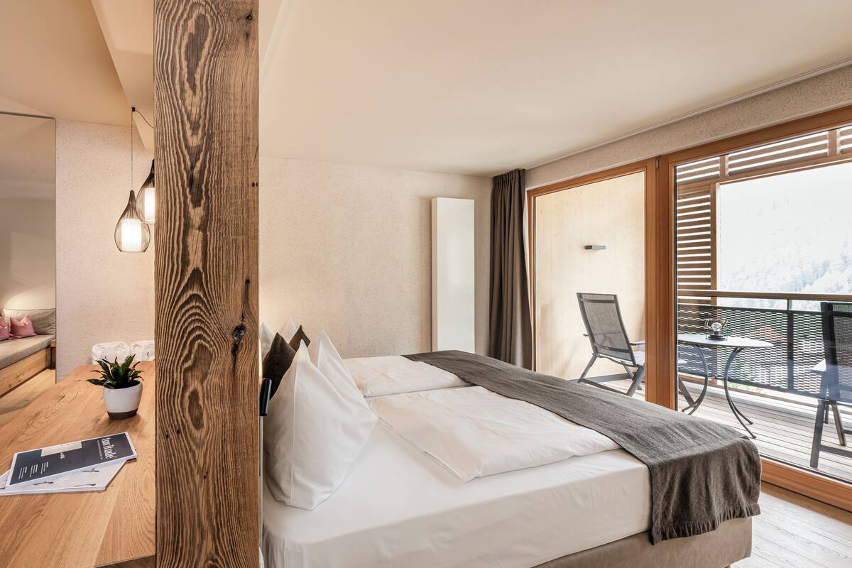 Ein modernes Hotelzimmer mit einer Wand aus Holzvertäfelung, einem Bett mit weißer Bettwäsche, einem kleinen Tisch mit Pflanze und einem Balkon mit Stuhl.