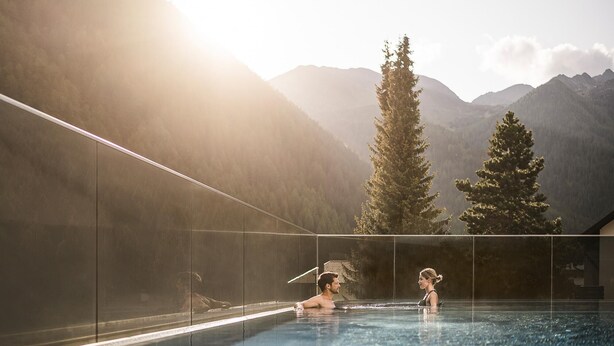 An infinity pool with a mountain backdrop, clear glass walls, and a person in the water. - Falkensteiner Hotel Falkensteinerhof  (Rio di Pusteria)