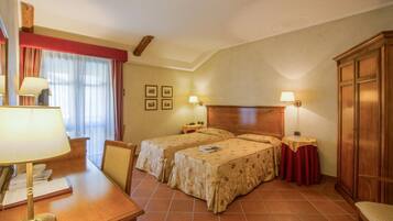 Habitación de hotel con dos camas, un escritorio con lámpara, una silla, un armario y una ventana con cortinas.