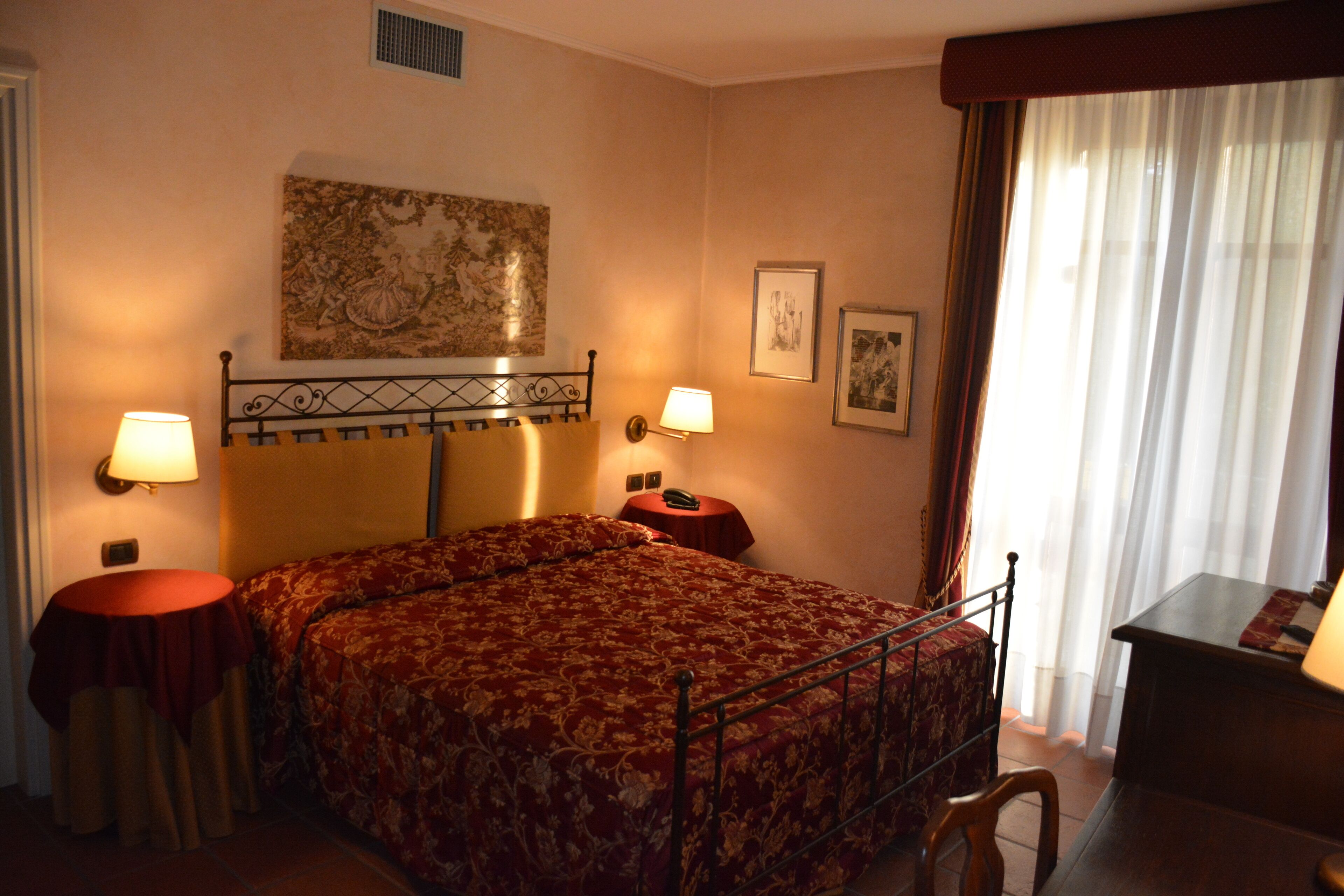A hotel room with a bed, two bedside tables, a lamp, a nightstand, a chair, and a window with curtains.