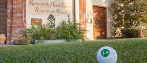 Une balle de golf sur l’herbe, devant un bâtiment sur lequel on peut lire l’inscription : « Romantik Hotel Furno Restaurant Relais ».