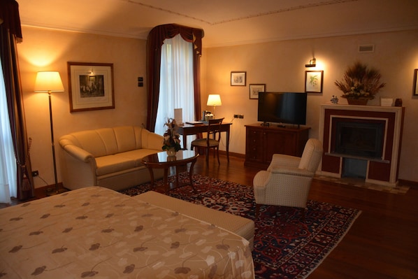 A hotel room with a bed, a sofa, a chair, a desk with a lamp, a television, and a fireplace.