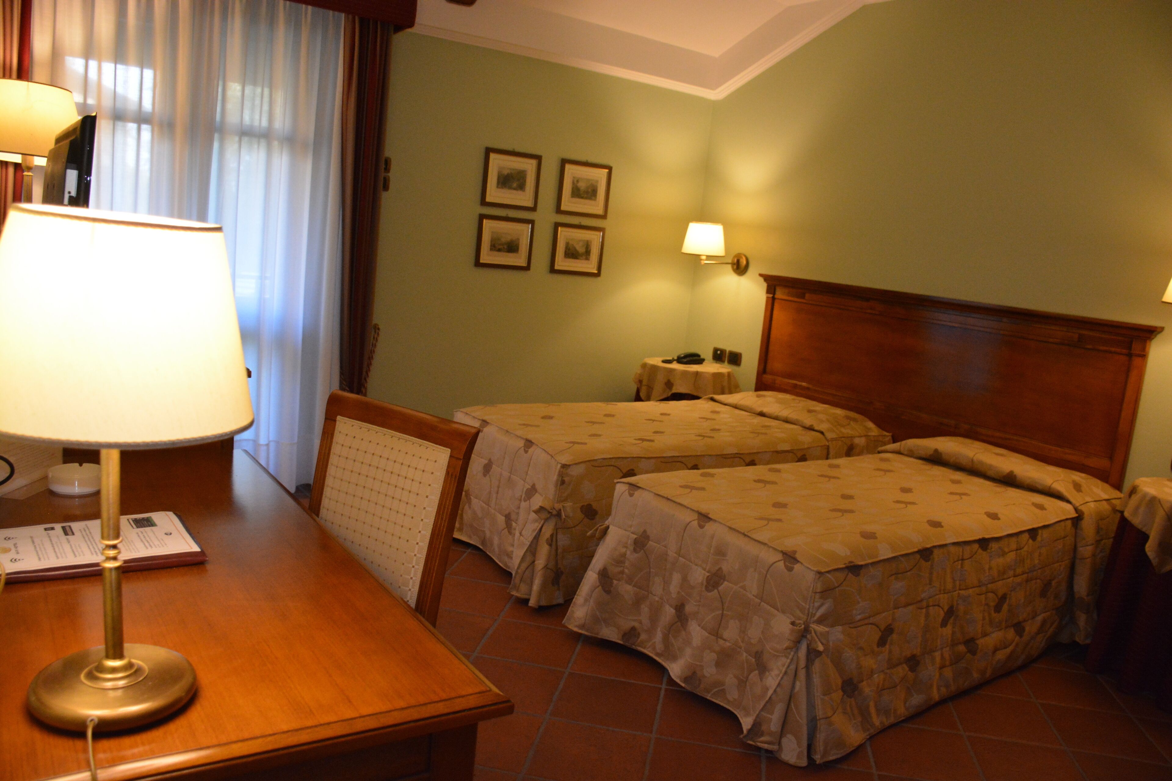 Habitación de hotel con dos camas, una mesita de noche de madera, un escritorio con lámpara, una pequeña mesa con lámpara y una ventana con cortinas.