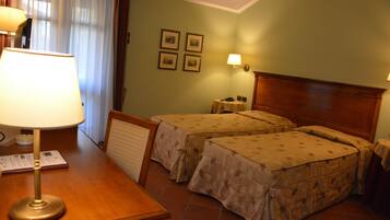 Habitación de hotel con dos camas, una mesita de noche de madera, un escritorio con lámpara, una pequeña mesa con lámpara y una ventana con cortinas.