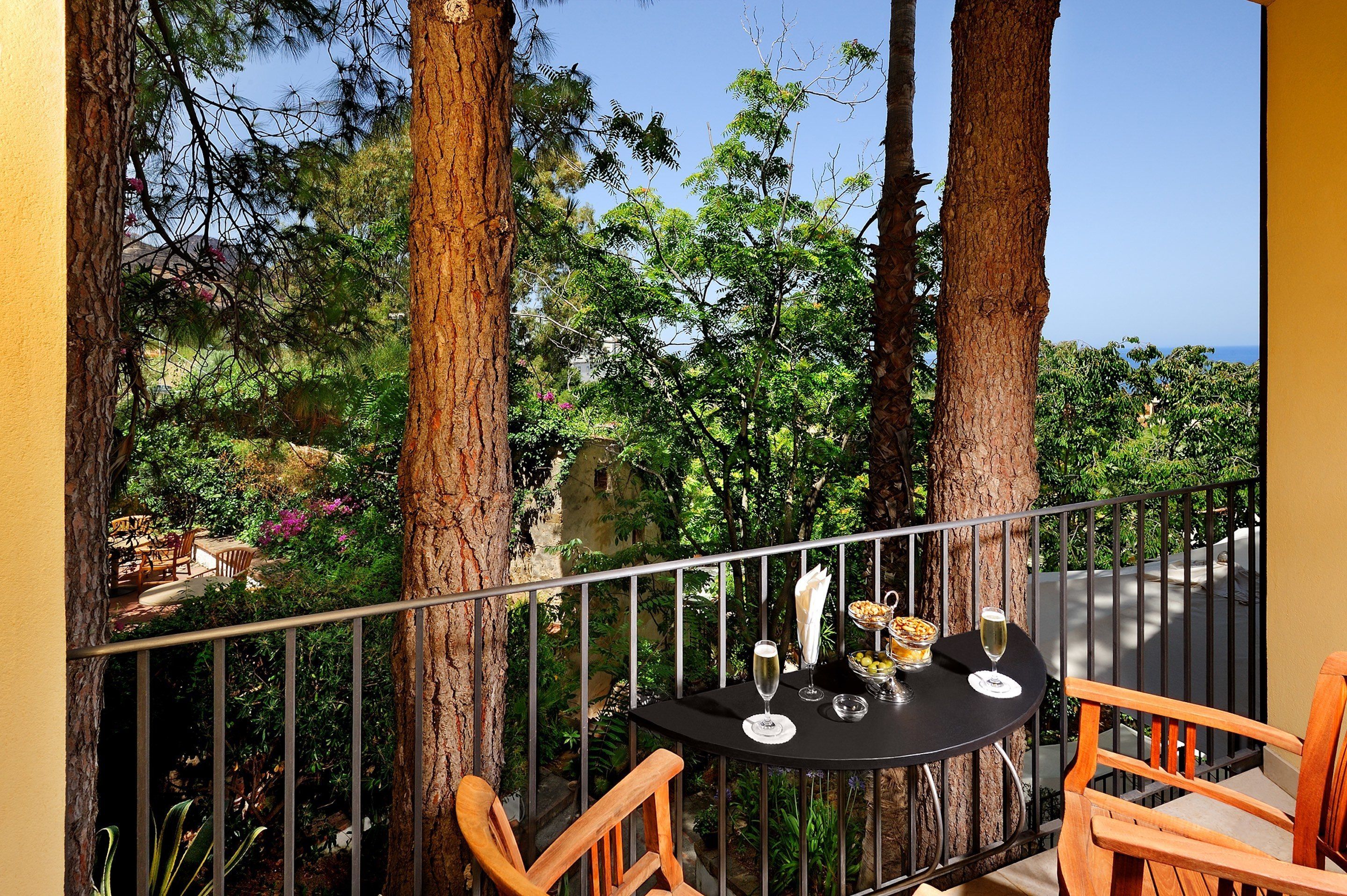 A balcony with a table set for two, surrounded by trees and a view of a lush garden and distant landscape.