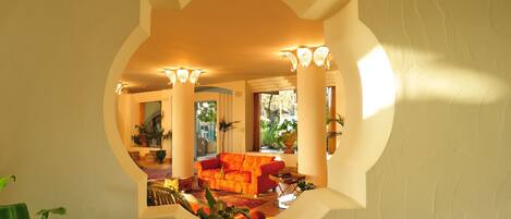 A room with a large mirror reflecting a cozy seating area, including a sofa and a coffee table. There are potted plants and a view of a cityscape through large windows.