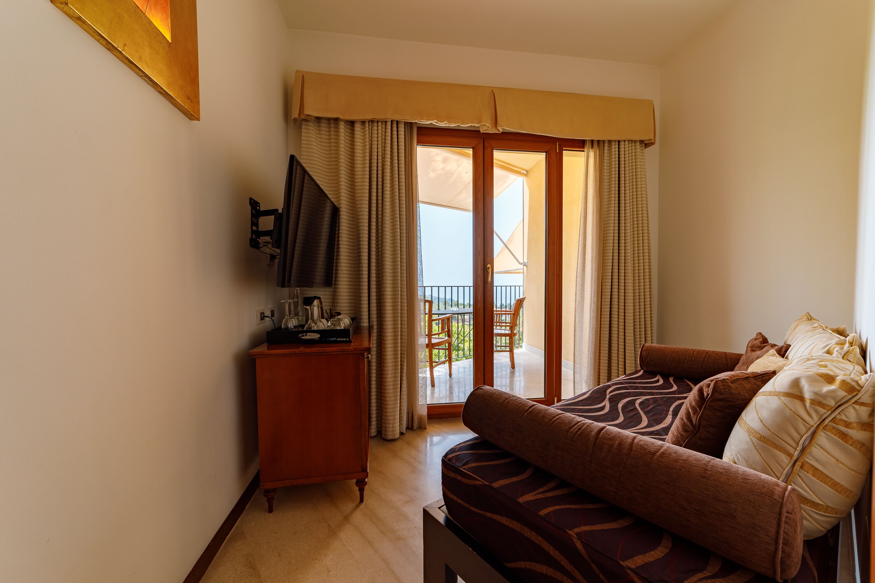 A hotel room with a sofa, a television, a small wooden cabinet, and a balcony with a table and chairs.
