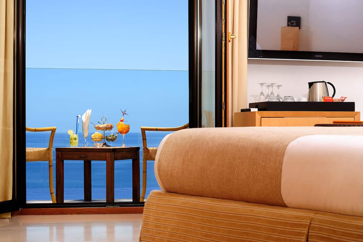 A hotel room with a large window overlooking the sea, a bed, a small table with a fruit bowl and glasses, and a shelf with a kettle and glasses.