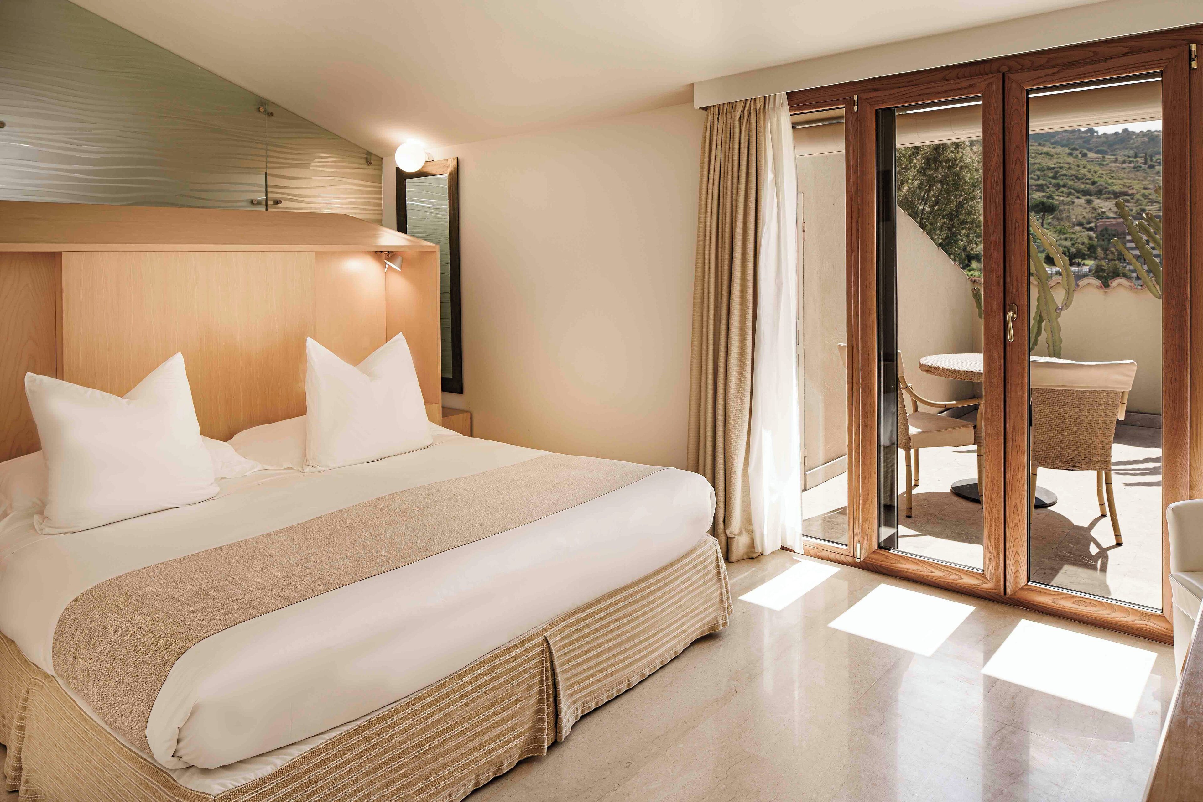 Habitación de hotel con una cama grande, cabecera de madera, balcón con mesa y sillas, y vistas al exterior.