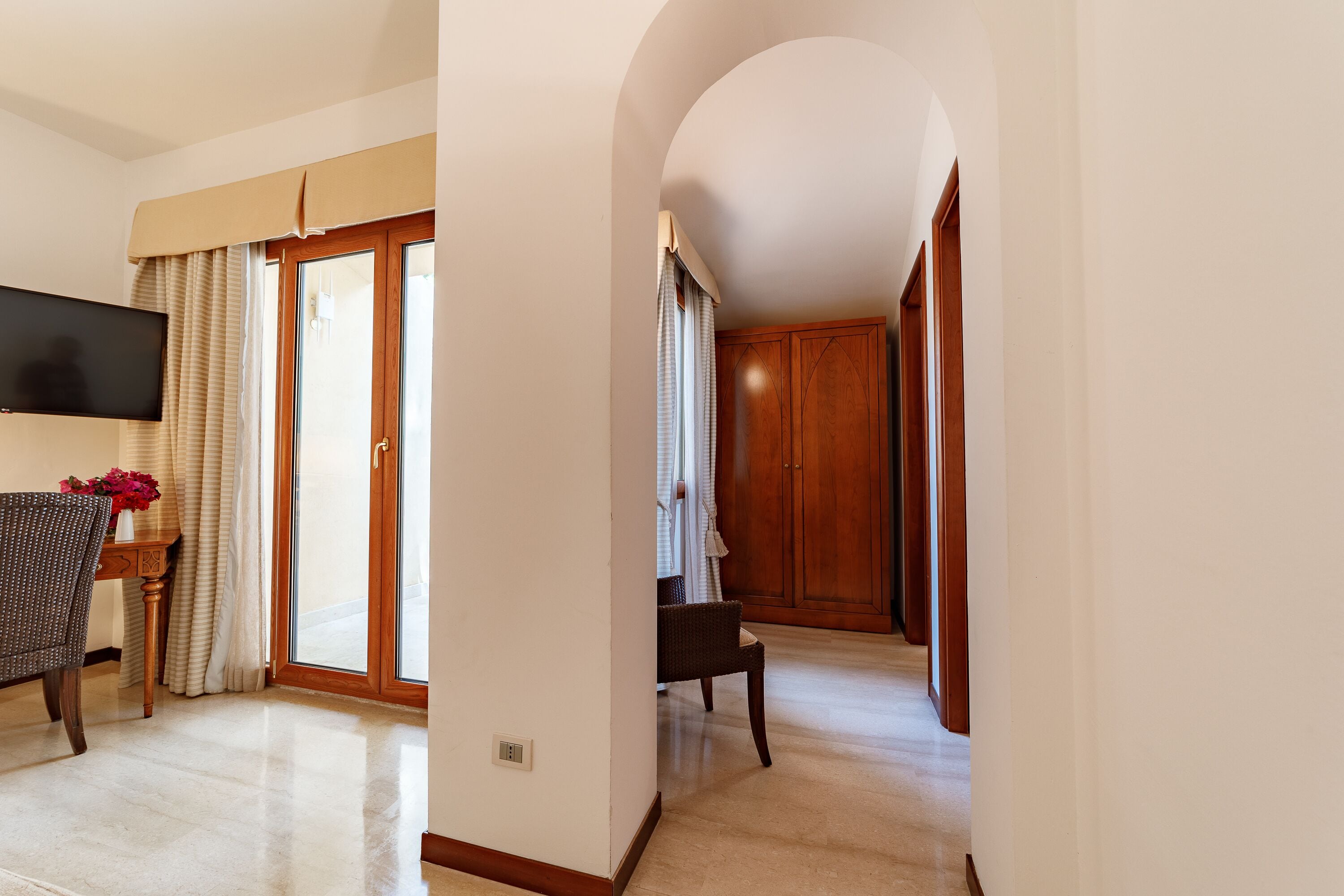 A hotel room with a flat-screen TV, a dining table with chairs, a wicker chair, a wooden cabinet, and a sliding glass door leading to a balcony.