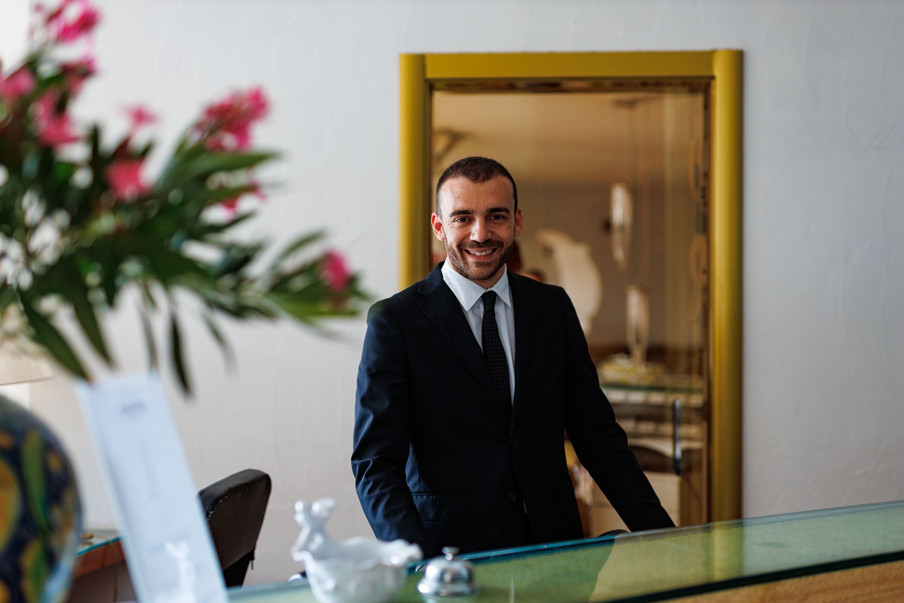 A man in a suit standing by a glass table with a chair and a plant in the background.