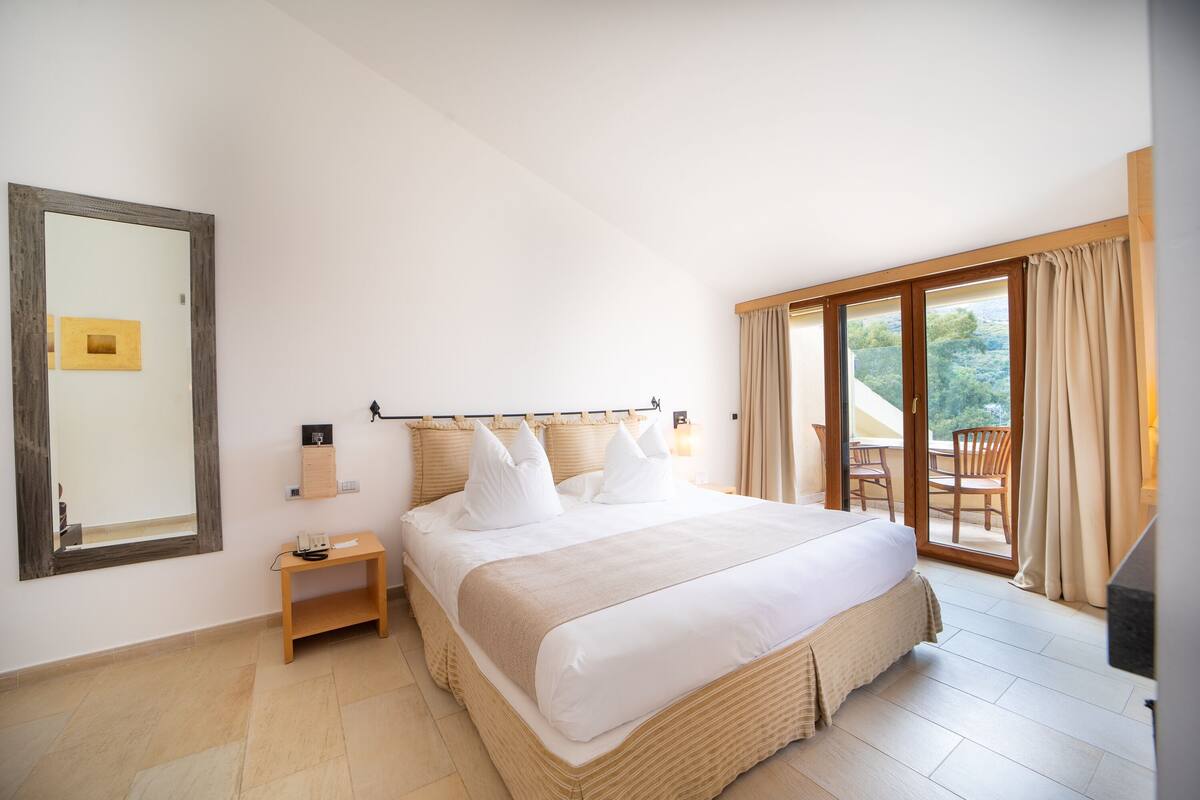 A spacious bedroom with a large bed, a bedside table, a mirror, and a view of the outdoors through a sliding glass door.