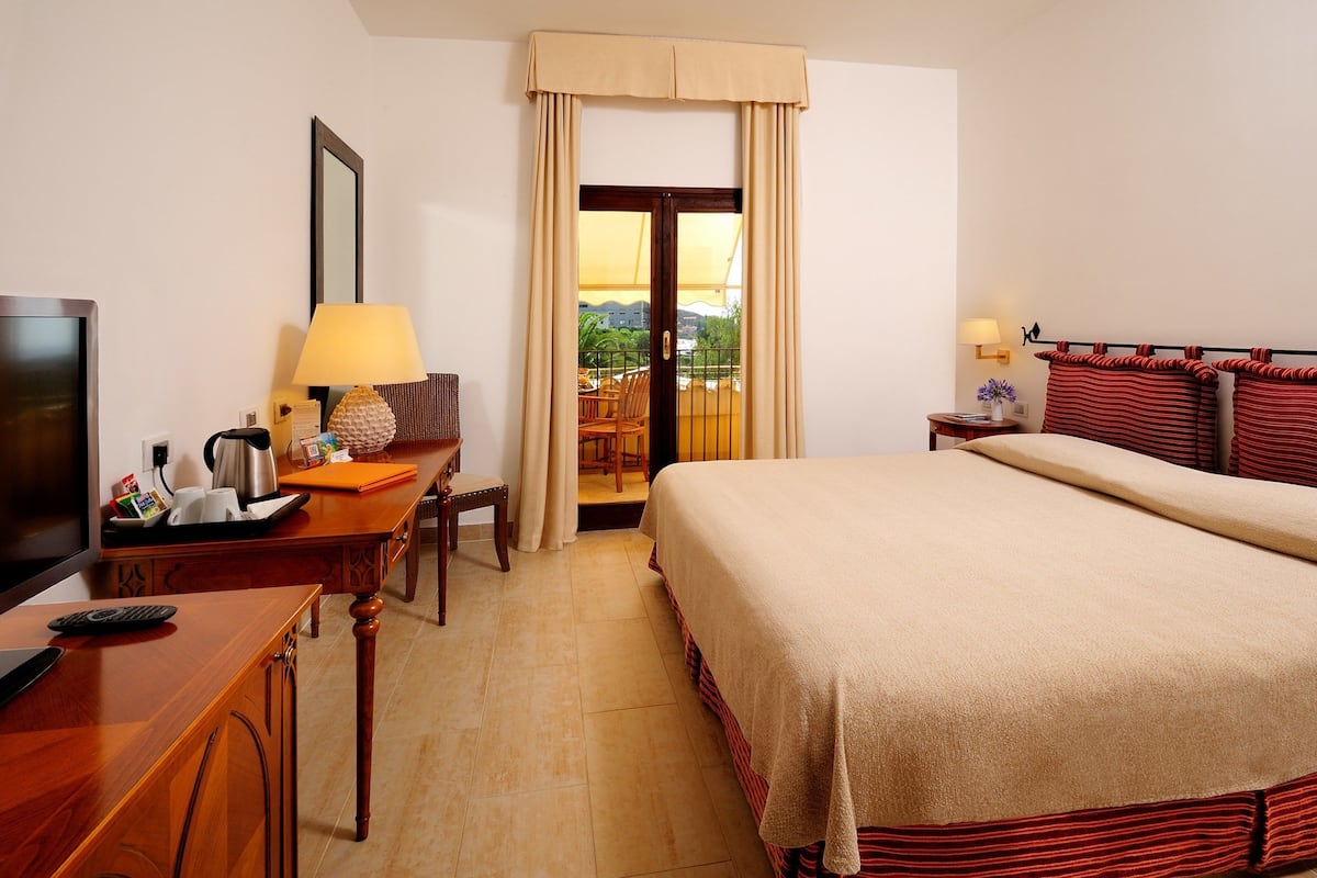 A hotel room with a bed, a desk with a lamp, a TV, and a view of the outdoors through a door.