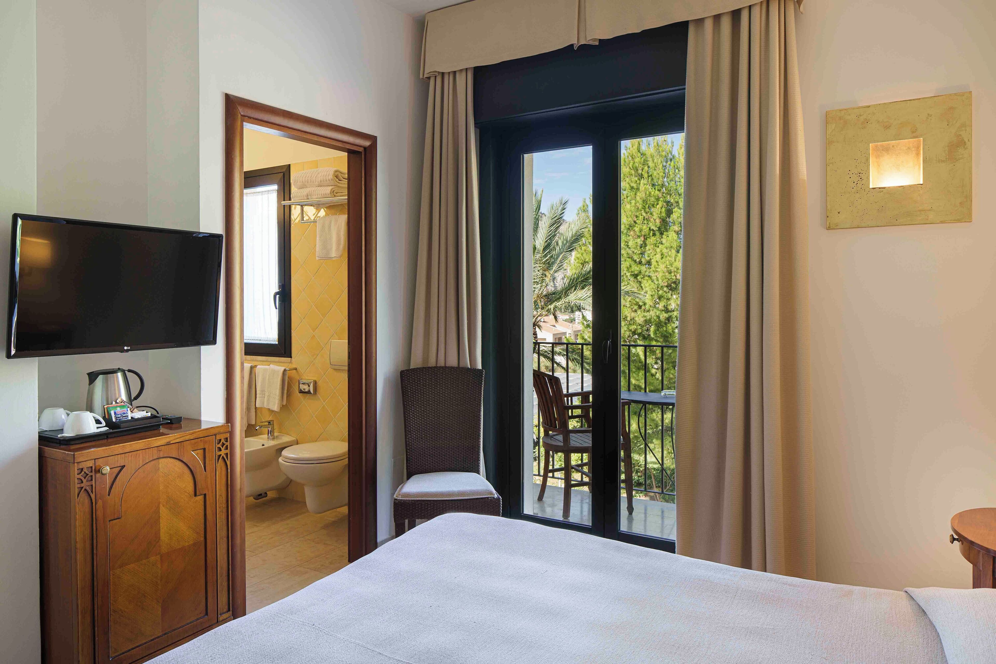 A hotel room with a bed, a TV, a small wooden cabinet, a chair, and a balcony with a view of greenery.