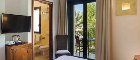 Habitación de hotel con cama, televisor, un pequeño mueble de madera, una silla y balcón con vistas a zonas verdes.