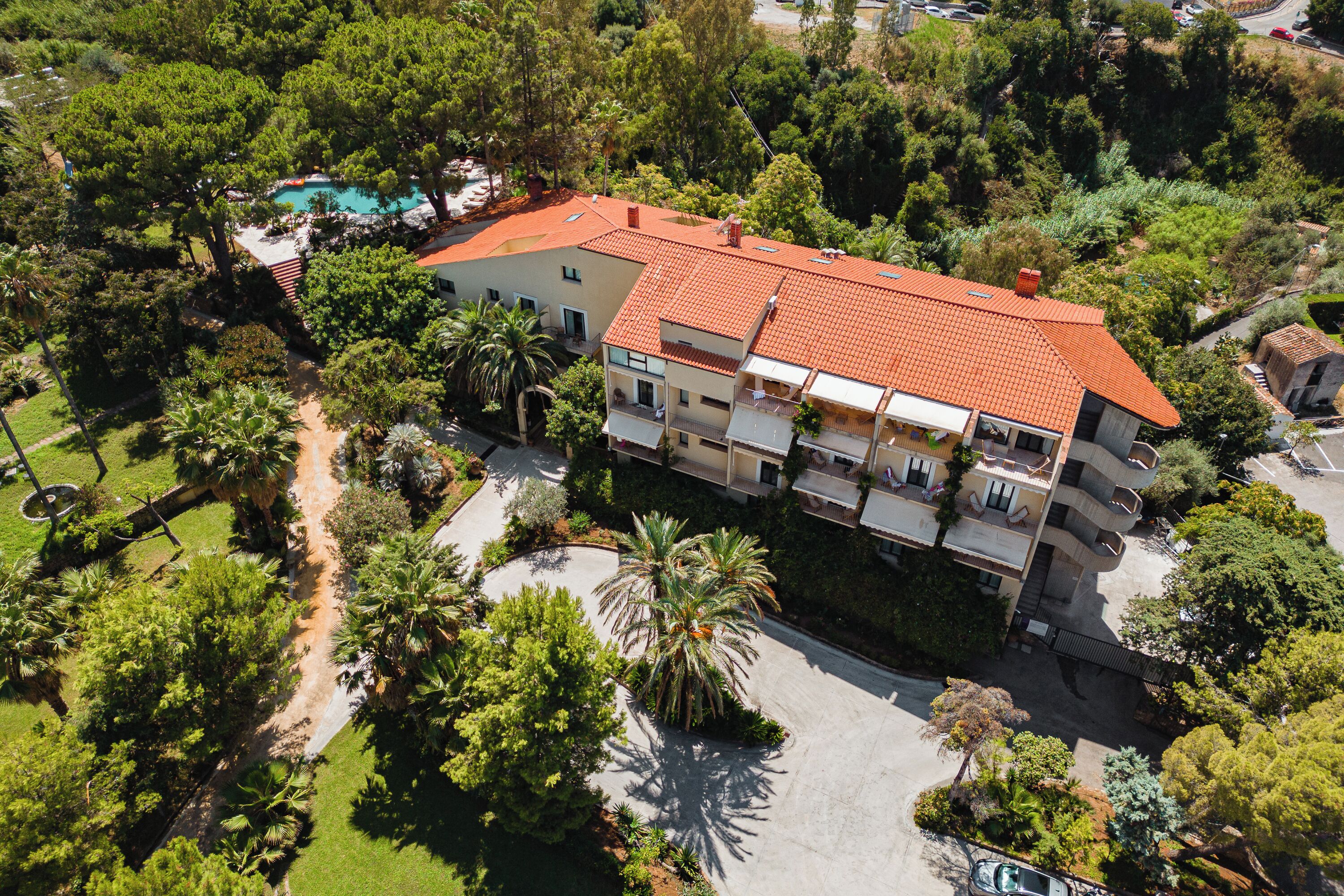 An aerial view of a multi-story building with a red-tiled roof, surrounded by lush greenery and a swimming pool.