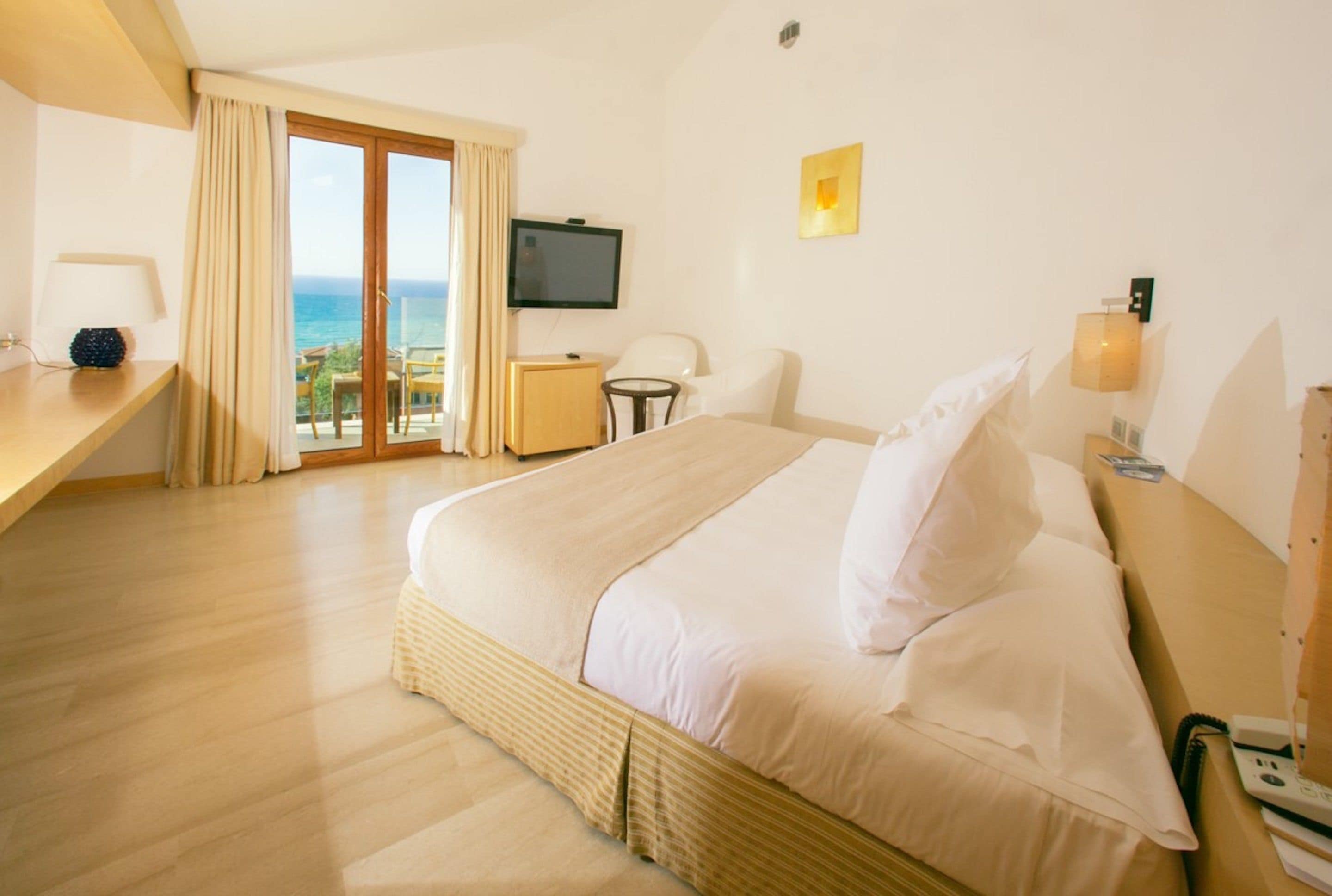 Habitación de hotel con cama, escritorio, silla, televisor y balcón con vista al mar.