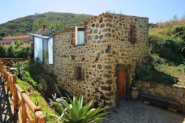 A stone building with a wooden door and window shutters, surrounded by plants and a wooden fence. - Hotel Alberi del Paradiso (Cefalù)